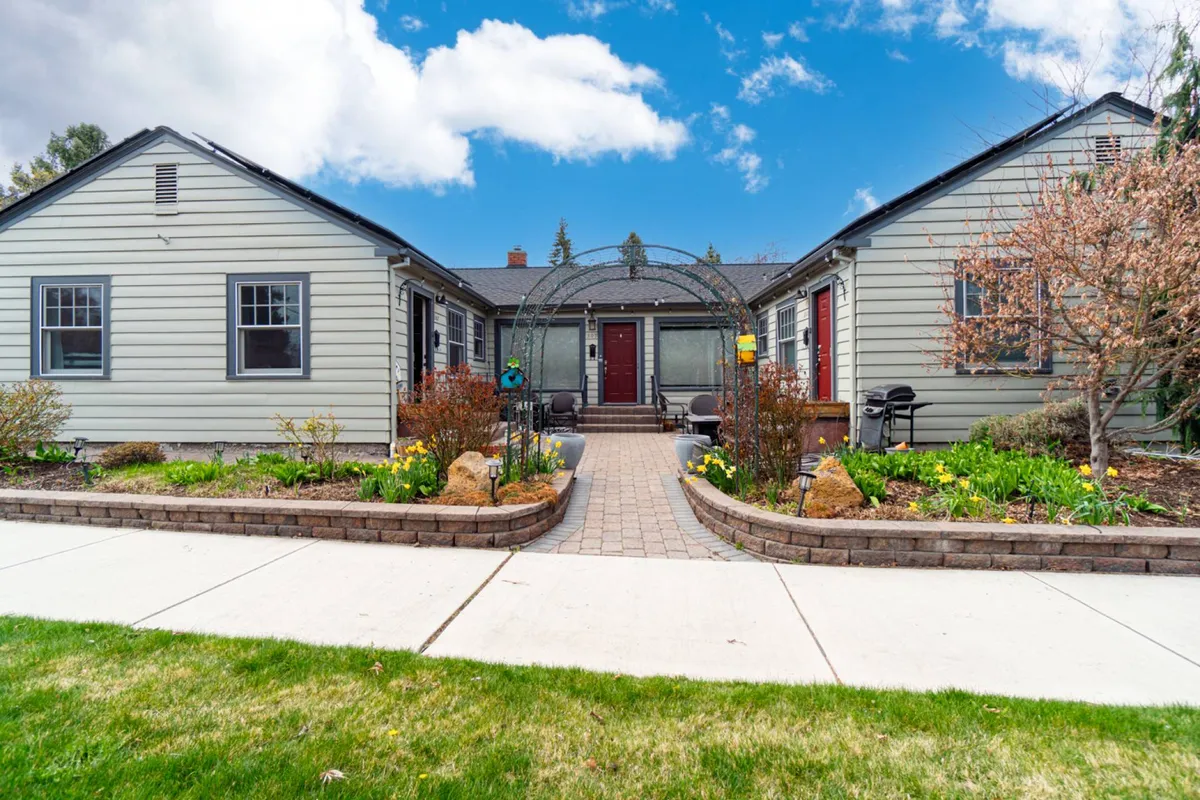The image showcases the charming exterior of a lovingly restored 1930s fourplex in downtown Bend, Oregon. An arched trellis frames the inviting front steps, leading to a cozy porch perfect for morning coffee. The well-maintained, landscaped garden adds a touch of tranquility, with greenery and flowers enhancing the cottage's curb appeal.