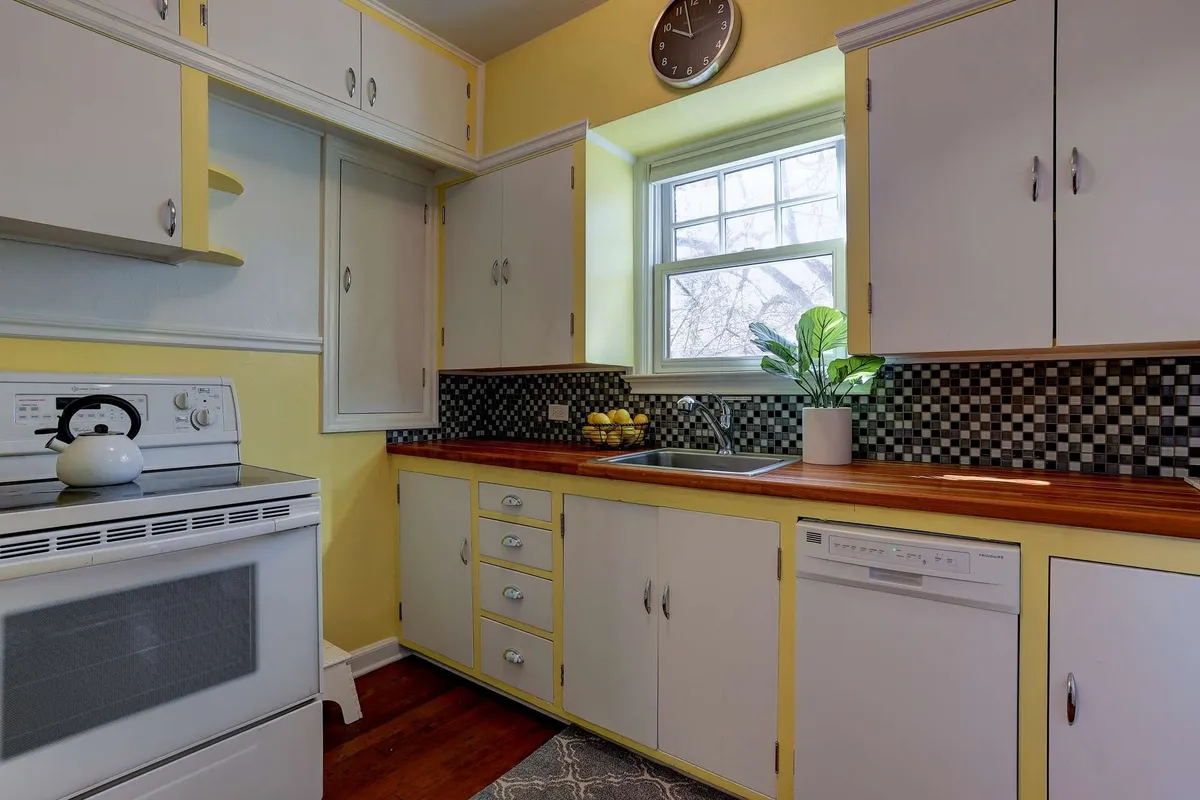 A bright kitchen corner featuring a well-lit sink under a window with a small plant, a checkered backsplash, a wooden countertop, and a clock. The scene suggests morning routines, such as brewing coffee, and conveys warmth and thoughtfulness in design.