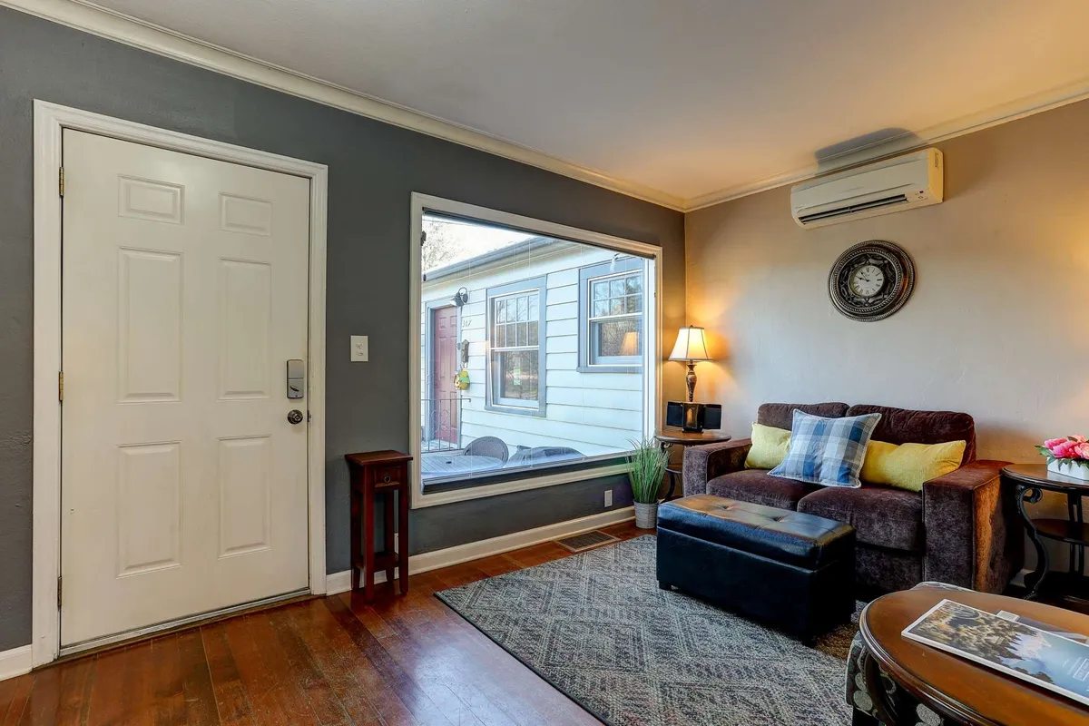 This interior image features the cottage's inviting front door area, a large window for natural light, a comfortable sofa, a table lamp, and a stylish rug. The space is bright, cozy, and conducive to both relaxation and work, hinting at the ease of transitioning between productivity and downtime.
