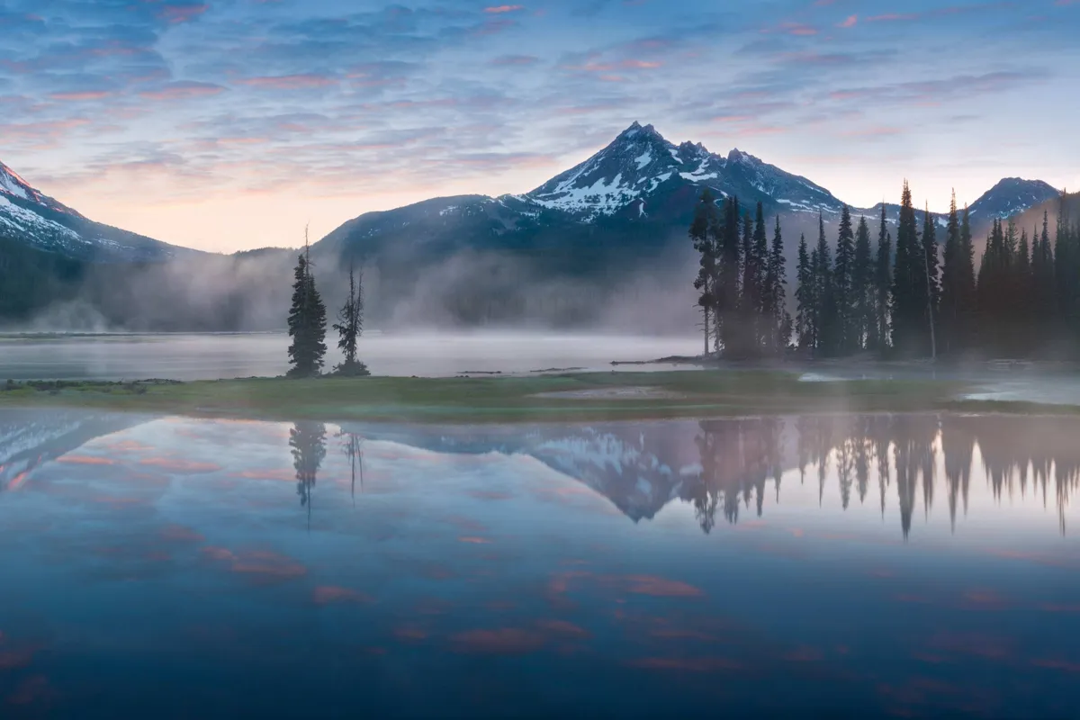 A serene lake bordered by pine trees, with majestic mountains rising in the background and a soft sky above. The environment is peaceful, inviting activities like hiking, birdwatching, or simply soaking in the view.