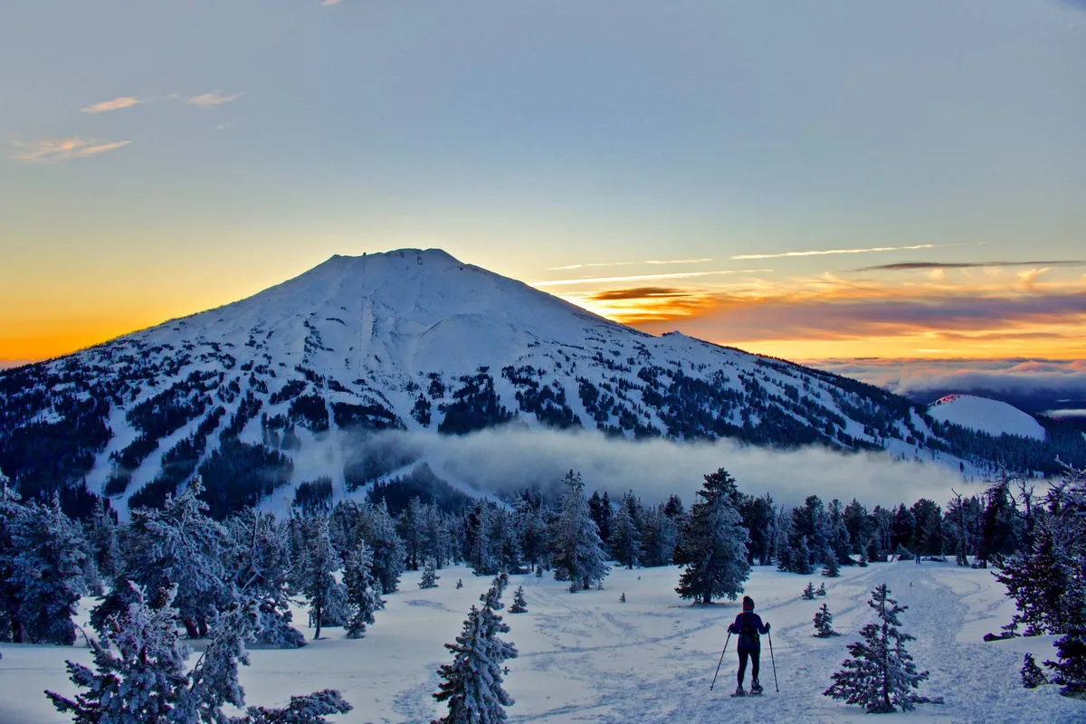 A breathtaking winter landscape featuring a snow-covered mountain, a lone hiker traversing the frosted terrain, and a dramatic sunset sky overhead. The scene captures the possibility of outdoor adventure and serene moments in nature.