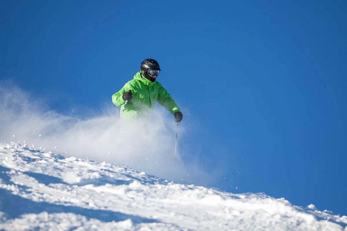 A skier in a bright green jacket carves down a pristine snowy slope beneath a vivid blue sky, with distant mountain peaks adding dramatic scenery. The image conveys the excitement of winter sports and the natural beauty accessible from Bend.