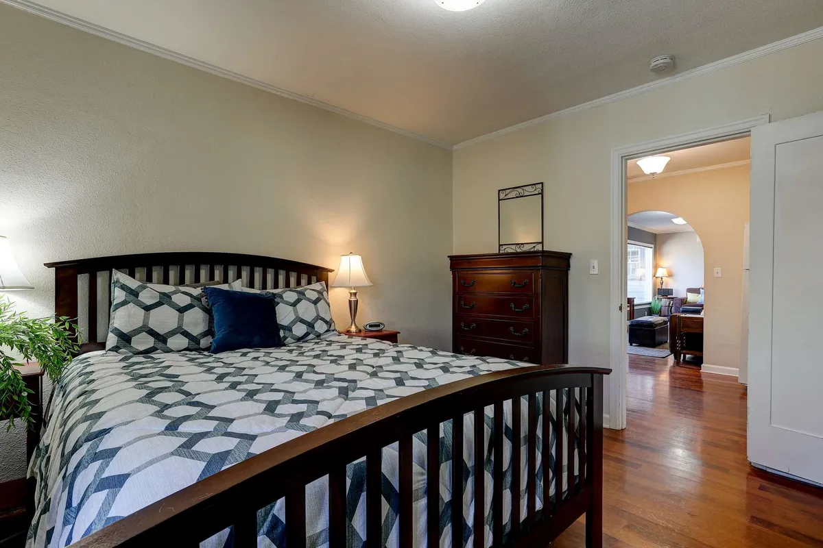 A cozy bedroom featuring a bed with geometric patterned bedding, a nightstand with a lamp, and a wooden dresser. Soft, warm lighting and minimal yet soothing decor create an inviting and restful atmosphere. The ample dresser and nightstand highlight practical storage features.