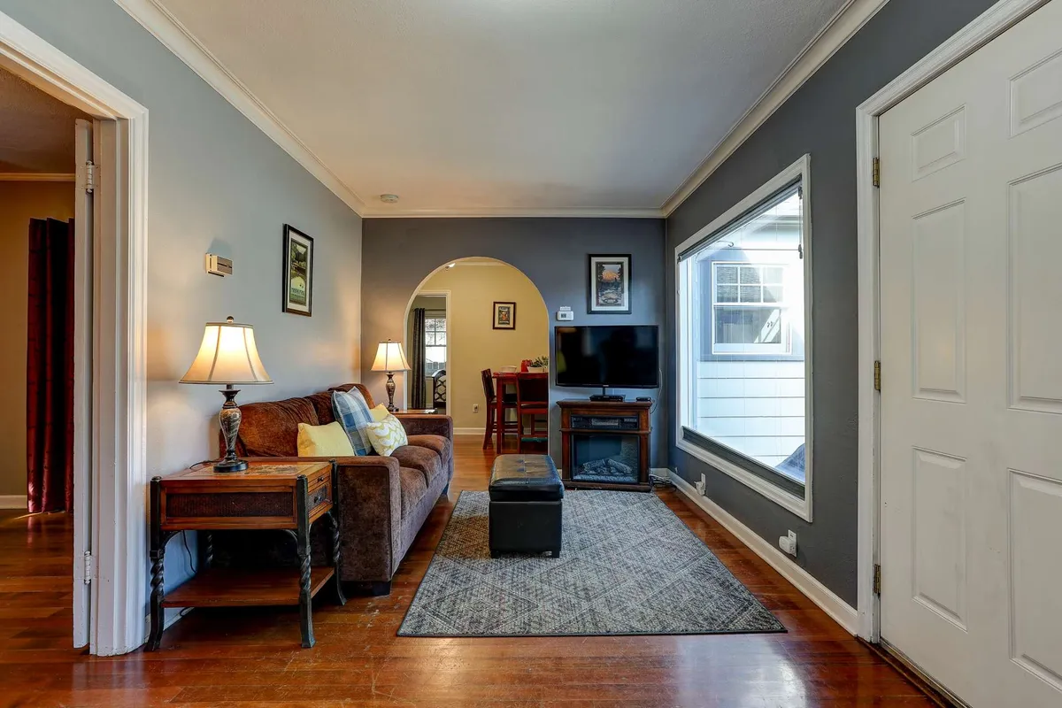A cozy living room featuring a comfortable sofa set around a coffee table, with a television, floor lamp, and a window letting in natural light. The decor blends vintage charm with contemporary touches, creating a space perfect for relaxing or socializing.