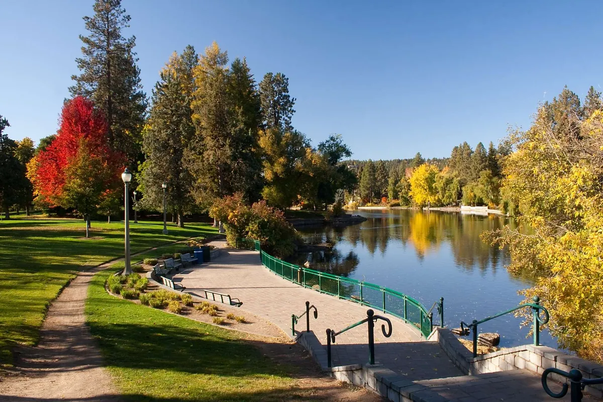 A tranquil view of a calm water surface bordered by colorful autumn trees, a walking path, and a green railing. The scene captures the essence of Drake Park, inviting visitors to enjoy peaceful moments by the water within the heart of downtown Bend.