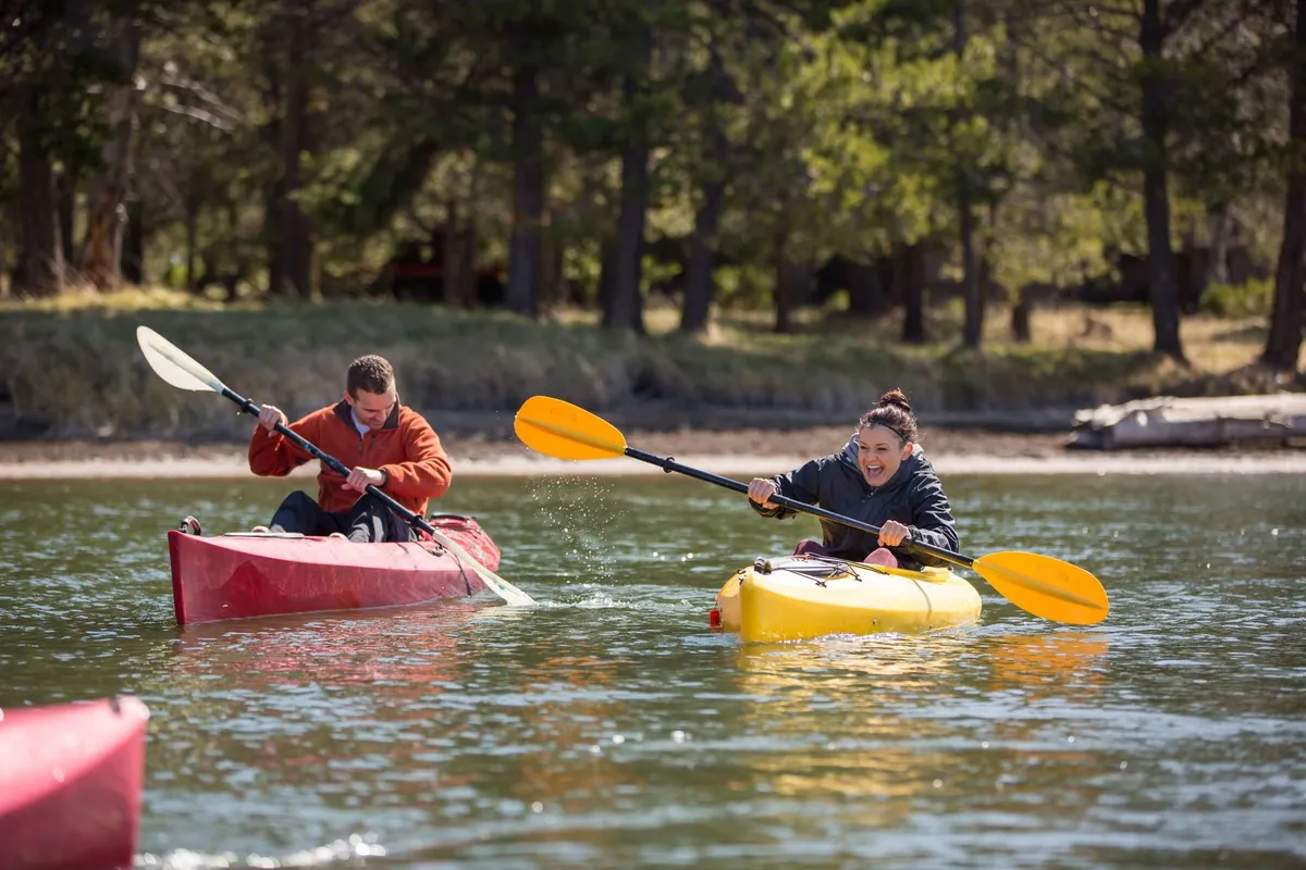 The scene shows two kayakers navigating a river framed by lush trees, capturing the adventurous spirit and access to outdoor recreation that defines Bend. The playful mood and connection to nature emphasize Bend’s blend of city energy and natural beauty.